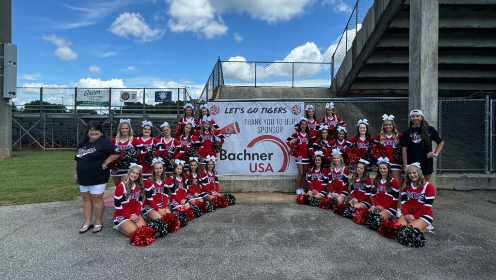 Cheerleaders standing and sitting in front of a poster, on which they thank Bachner for being a sponsor.